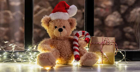 Wait for Santa. Teddy bear sitting on a window sill, Christmas lights and gift box, warm festive home interiorの写真素材