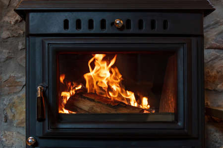 Wood stove fireplace and burning firewood behind glass door, Black metal fireside, fire flame radiate heat, warm home interior,の写真素材