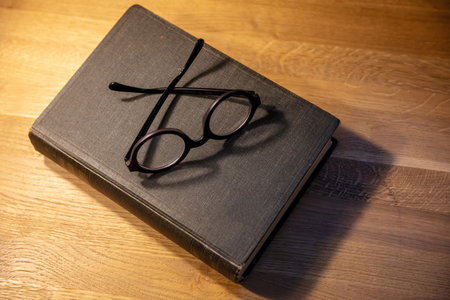 Closed old black book on a table. Hardcover notebook and eyeglasses on wooden background, top view....