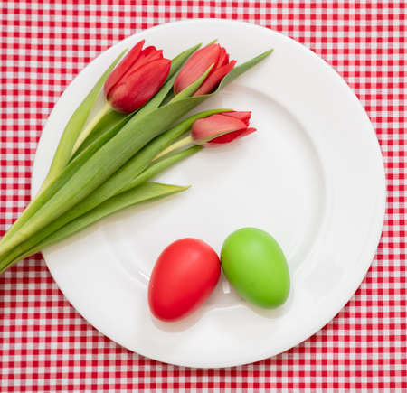 Easter Holiday table. Egg red and green and tulips decoration on white plate, checkered tablecloth background, top view,の写真素材