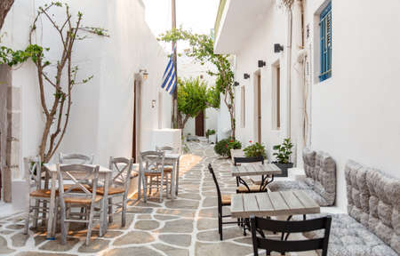 Paros island outdoor cafe tavern at Naousa Greece. Traditional whitewashed building plant empty wooden table and chair on narrow cobblestone alley summer day.の写真素材