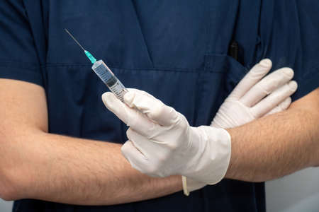 Doctor in medical blue uniform holding in hand with disposable glove, liquid drug in syringe with...