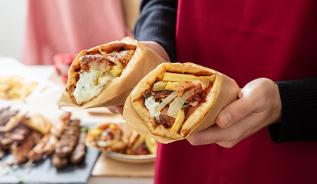 Gyro pita Shawarma.  Greek food, sliced meat, potato, tomato and tzatziki, paper wrap in male hand, kitchen background, close up view.の写真素材