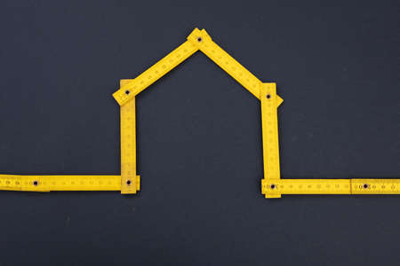 Home repair. House shape yellow wooden measure, gray black background. Folding ruler, workshop, diy, handyman concept.の写真素材