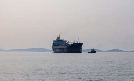 Small tugboat, powerful watercraft tows cargo ship freighter to Ermoupoli city port, Syros island,...