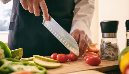 Chef with black apron cutting tomato on a chopping board, cook hand holding a knife close up. Fresh...
