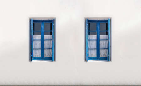 Two wooden windows with blue open shutters on white wall background. Cyclades island house front view, Greek traditional architecture.の写真素材