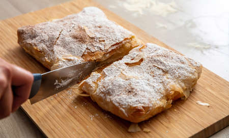 Greek pastry with cream sugar powder and ground cinnamon. Hand with a knife cutting bougatsa,...