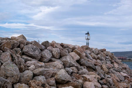 Lighthouse on seawall, protection at navigation, cloudy sky. Beacon on breakwater, close up view of rocky wall construction at harbor entrance of Greek island.の写真素材