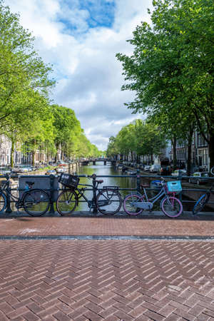 Bicycles parked on a bridge tiled floor over canal, moored boats on water. Amsterdam, Netherlands. Traditional houses brick facade and treesの写真素材