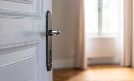 Retro door knob on white vintage wooden door, close up view. Door open, blur elegant house room interior, window with curtains and wood floor parquetの写真素材