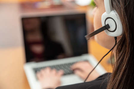 Call center, home office, customer support, help desk concept. Woman with headset working with a laptop, talking on phone, view from behindの写真素材