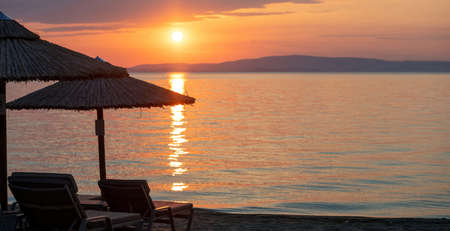 Sunset on the beach over Aegean Sea, Greece. Straw umbrellas and sun beds on the sand, reflections on ocean water, orange color sky and cloudsの写真素材