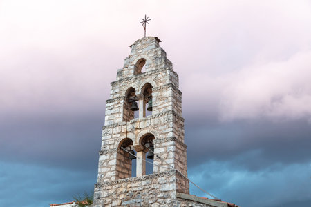 Belfry with four metal bronze bells at Mani Laconia, Peloponnese Greece. Under view of traditional stonewall bell tower with cross, cloudy sky background. Orthodox religious destination.の写真素材