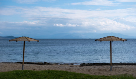 Two wooden parasol on an empty sandy beach. Winter day, blue rippled sea and cloudy sky background. Seascape from Greece.の写真素材