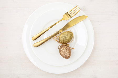Easter table setting, Golden Easter eggs decoration, napkin and gold cutlery on plates, white wooden...