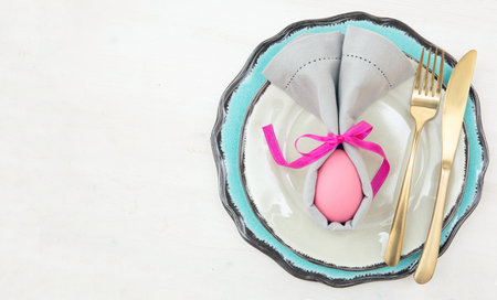 Easter table setting and decoration, Easter egg in a gray napkin, golden cutlery on plates set,...