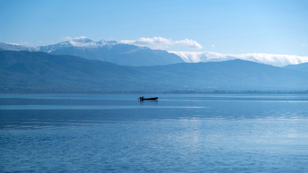 Greece Pamvotis Lake, Ioannina city Epirus. All shades of blue on calm water and clear sky background. Silhouette of fishing boat, snowy mountain peak.の写真素材