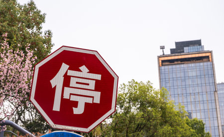 Stop road sign in chinese language. Red octagonal traffic sign for cars in China, cloudy sky, high rise building and blossoming pink flowersの写真素材