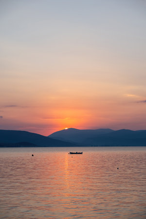 Tranquil fishing boat at sunset over Tolo Bay Greece. Glowing orange sky and distant mountain silhouettes reflecting on calm sea water surfaceの写真素材