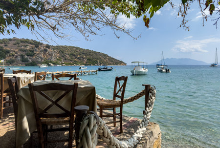 Charming seaside taverna with wooden chairs and tables overlooking anchored boats in the calm water of the Saronic Gulf, Greeceの写真素材