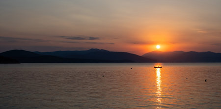 Tranquil fishing boat at sunset over Tolo Bay Greece. Glowing orange sky and distant mountain silhouettes reflecting on calm sea water surfaceの写真素材