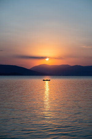 Tranquil fishing boat at sunset over Tolo Bay Greece. Glowing orange sky and distant mountain silhouettes reflecting on calm sea water surfaceの写真素材