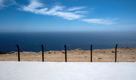 View of the vast blue sea framed by the wire fence over a white painted wall, blue sky with scattered clouds, evoking calm isolation. Gavdos island, Greeceの写真素材