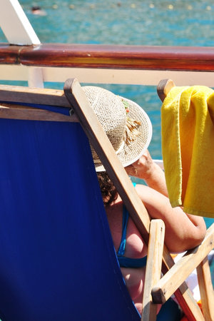 Rear view of adult woman wearing straw hat sitting on chaise longue and sunbathing on beach in summer dayの写真素材
