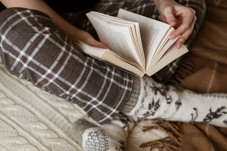 Woman in warm woolen knitted socks sits on sofa reading book, winter hygge leisure time
