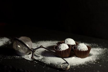 Chocolate candies sprinkled with powdered sugar and metal strainer lying on dark table surface, cook at home, homemade foodの写真素材