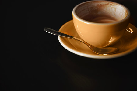 Half empty yellow coffee cup on plate with spoon on dark background