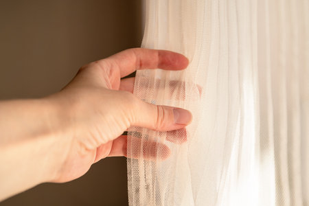 Woman adjusts ivory colored tulle with her hand, window light, close-up handsの写真素材
