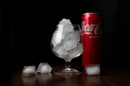 Wine glass filled with ice cubes with red defocused cola can on dark background