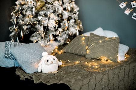 Festive home interior with decorative cushions scattered on the floor under ornate decorated Christmas tree, lighted garland and cute toy on the couchの写真素材