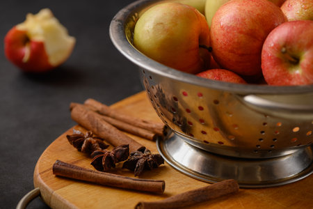 Metallic bowl full of red ripe apples on wooden cutting board with scattered cinnamon sticks with...