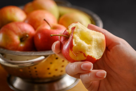 Bitten apple in human hand on metallic bowl full of red ripe apples background. Healthy eating. Apple pie ingridients. Cooking at homeの写真素材