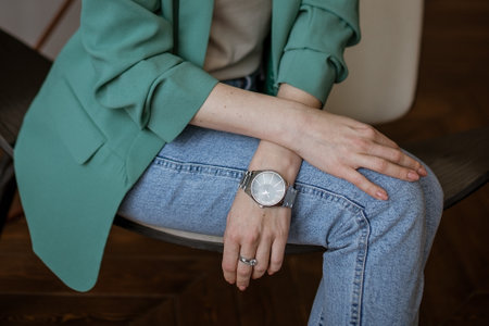 Pretty woman in blue jeans and green jacket sits with her hand on her lap,  stylish watch on her...