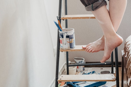 Woman with bare crossed feet stained with white paint stands on stepladder. Home renovation conceptの写真素材