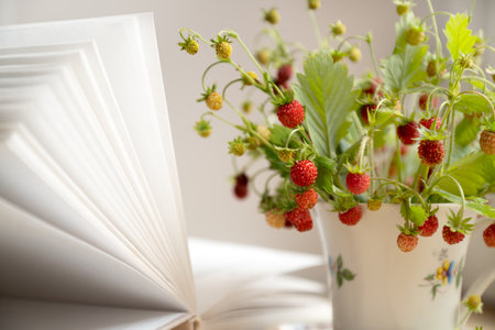 ripe red berries of wild strawberries on  twigs  collected in bunch in tea cup next to open bookの写真素材