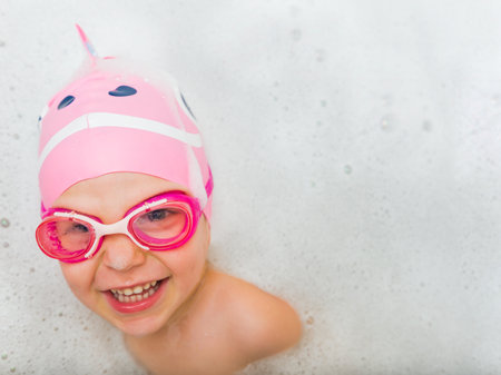 A happy child wearing goggles and a fun swim cap, enjoying swimming surrounded by foam bubbles.の写真素材