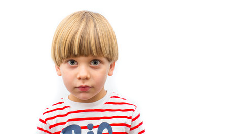 A front view of a young boy with blond hair, wearing a striped T-shirt, looking thoughtful while posing against a bright, neutral background. The simple attire and expression add a touch of innocence.の写真素材