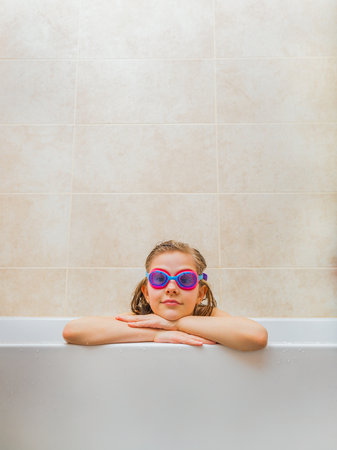 Bright and cheerful image of a young girl enjoying a bath while wearing pink swim goggles. The setting is clean, modern, and highlights a playful, childlike spirit with a focus on water-related themes.の写真素材