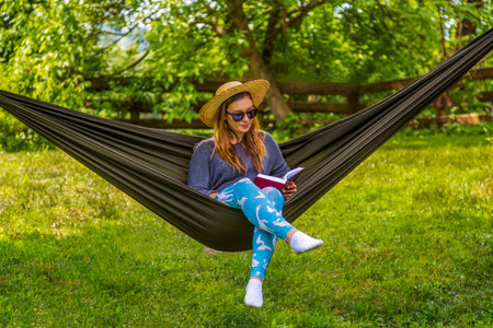 A young woman lounges in a hammock, surrounded by lush greenery, enjoying reading a book during a sunny, relaxing day in a bright outdoor environment.の写真素材
