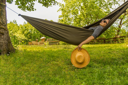 A serene view of a woman reclining in a hammock, surrounded by lush greenery and basking in the summer sun, holding a straw hat in a relaxing outdoor setting.の写真素材