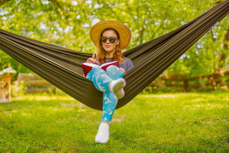 A woman enjoys a relaxing moment in nature, lounging in a hammock while reading a book. Surrounded by lush greenery, she embraces the tranquility and warmth of a sunny day.の写真素材
