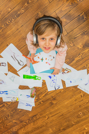A cheerful young girl with headphones is surrounded by her creative artwork while sitting on a wooden floor.の写真素材