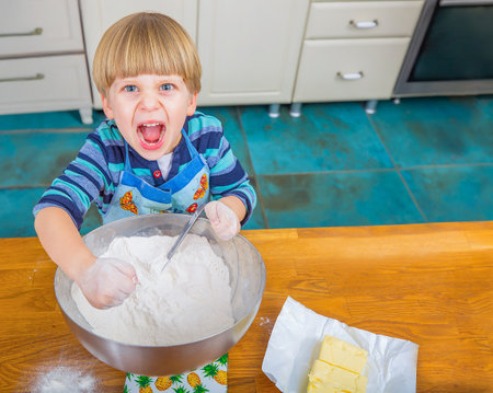 Energetic boy mixing ingredients for baking, expressing joy and excitement, creating a lively atmosphere in a kitchen with wooden countertops and tiled flooring.の写真素材
