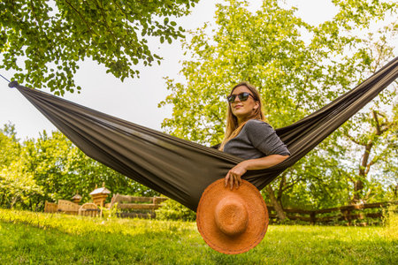 A confident woman enjoying the outdoors while relaxing in a hammock with a straw hat in hand. The serene garden setting showcases lush greenery and vibrant summer vibes, perfect for tranquility.の写真素材