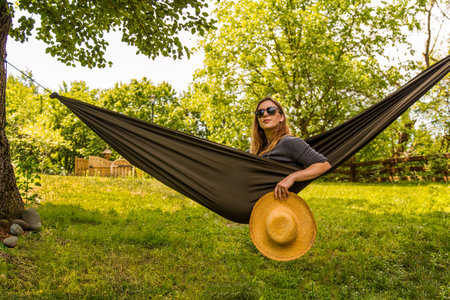 A woman relaxes in a hammock, holding a straw hat, surrounded by a tranquil green garden. The scene evokes calmness and connection with nature.の写真素材